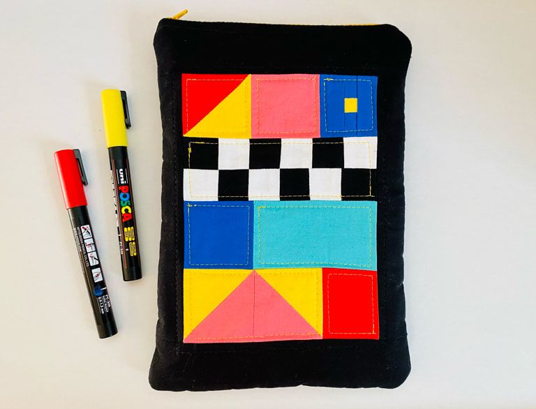 A black pouch with a patchwork panel made of triangles, squares, and rectangles. Colors include red, yellow, pink, blue, teal, black, and white. There are black and white squares in a checkboard pattern. Two red and yellow Posca markers are next to the pouch