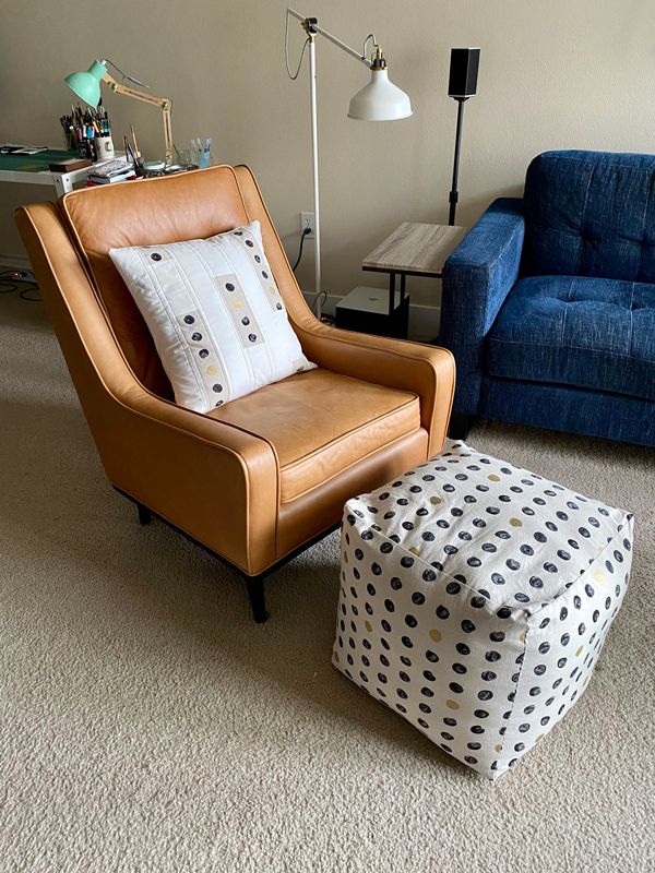 A living room scene shows a brown leather chair with a pillow on top, and a poufy cube ottoman in front of it. The pouf is made out of a natural linen with a design of large painted black-and-gold dots, and the pillow has strips of this and cream fabric.