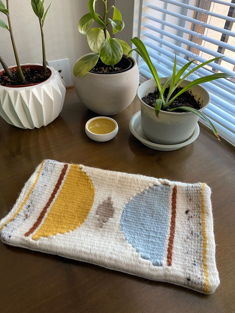 A poofy, large woven mug rug lying on a table. Two half circles in goldenrod and light blue point towards a tawny diamond in the center. The backdrop is a cream yarn, and there are also bands of speckled grey, goldenrod yellow, and brick red. There’s little gaps in the weaving on the edges of the half circles.