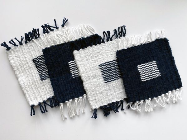 A set of square coasters with tassles. In the middle of each coaster is a square made up of alternating squiggles in contrast to the main color (either navy or white)