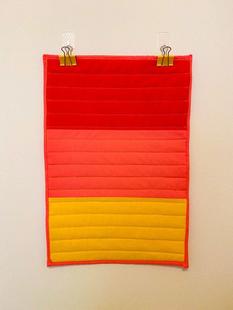 An 11 x 17 wall hanging with 3 thick bands of red, salmon pink, and yellow cotton fabric. The binding is salmon pink. There is a pattern of horizontal quilted lines 1.5 inches apart.