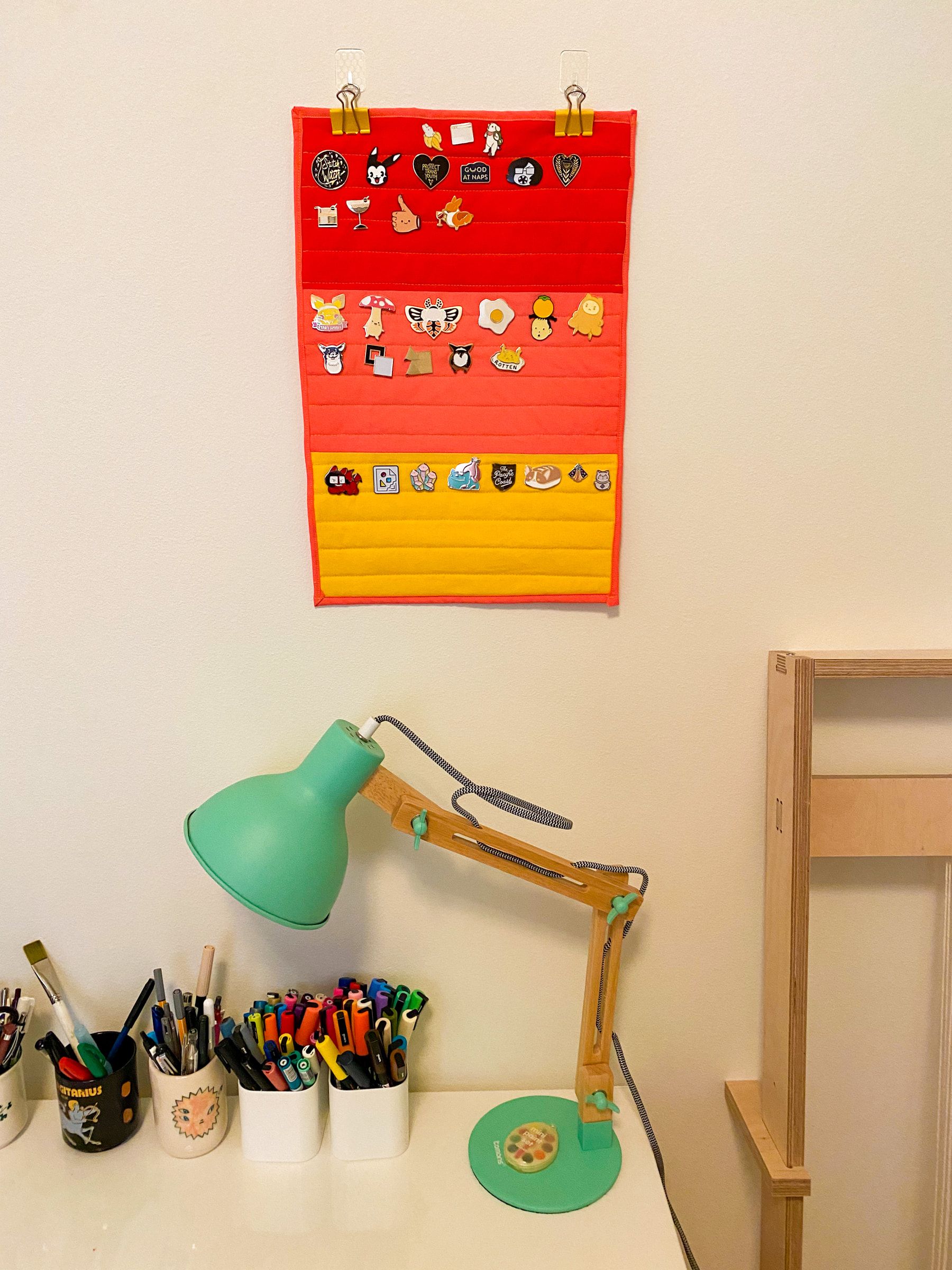 The banner hanging over my desk, which is white metal with a teal lamp on it. There's pen cups full of pens and markers.