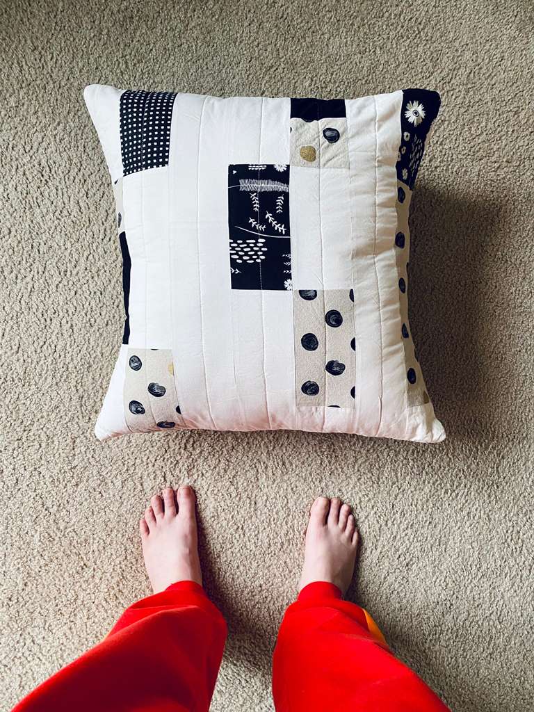 The pillow is thrown on a carpeted floor, and in front are my feet and legs in bright red sweatpants. The pillow is square and about three foot-widths in either direction.