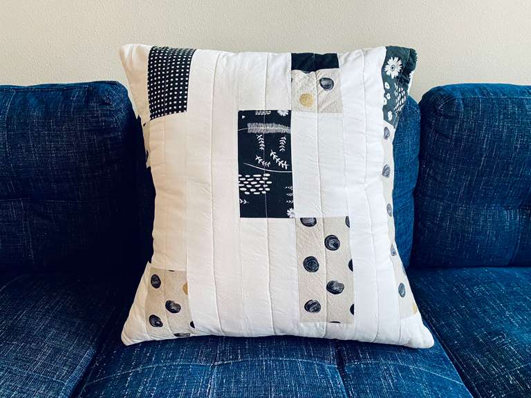 An overstuffed 24” quilted pillow made from randomly placed strips of fabric, sitting on a dark blue couch. The fabric is; black and white floral, black with white dots, cream with black and gold dots, and solid pale cream.