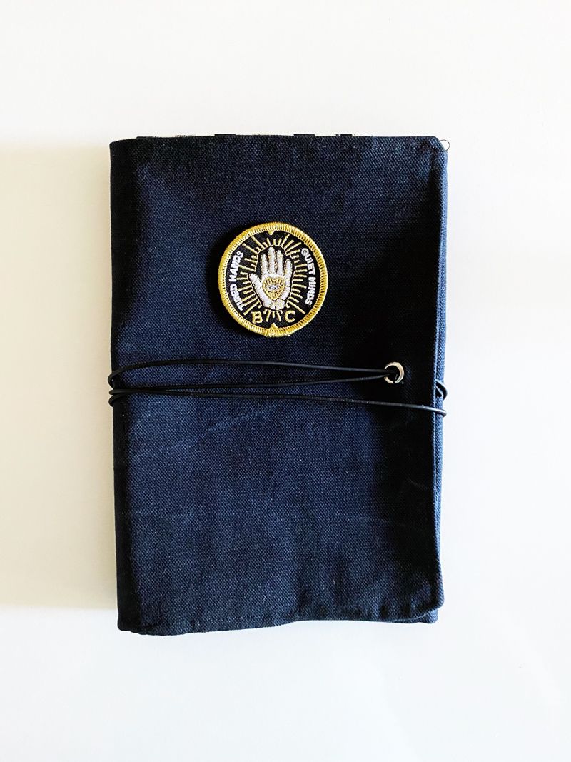 A folded, black canvas tool roll with a black leather cord wrapped around it. A patch on the front reads 'tired hands, quiet minds' and has a glowing hand illustration on it.