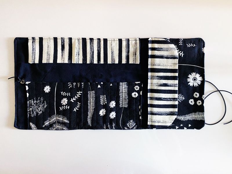 The tool roll unfolded. There is a pocket with a flap, and a row of spaces for pencils to slide in (also with protective flap). Materials used are a black and white floral print, and a painterly striped black and white print.
