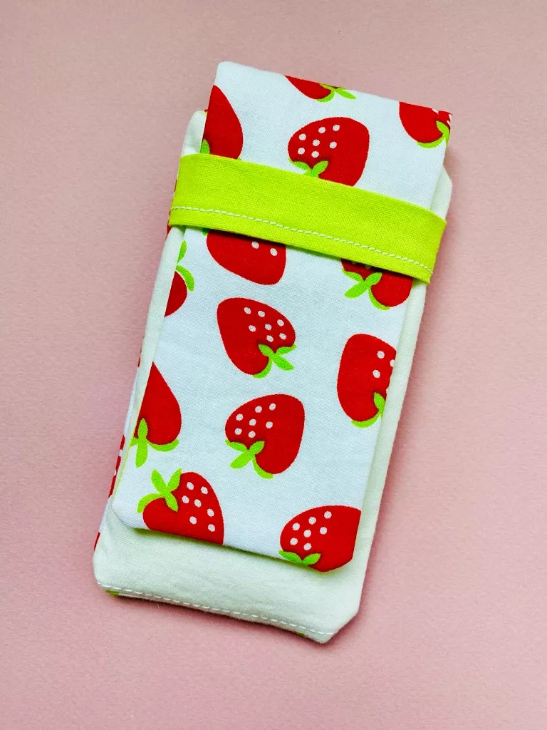 A fabric pen pouch featuring a strawberry pattern on the flap and a bright green strap that it tucks into.